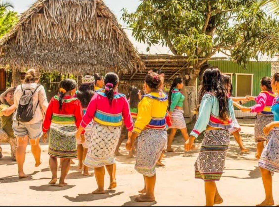 Group of people in colorful traditional clothing dancing outdoors near a thatched hut in sunny weather.